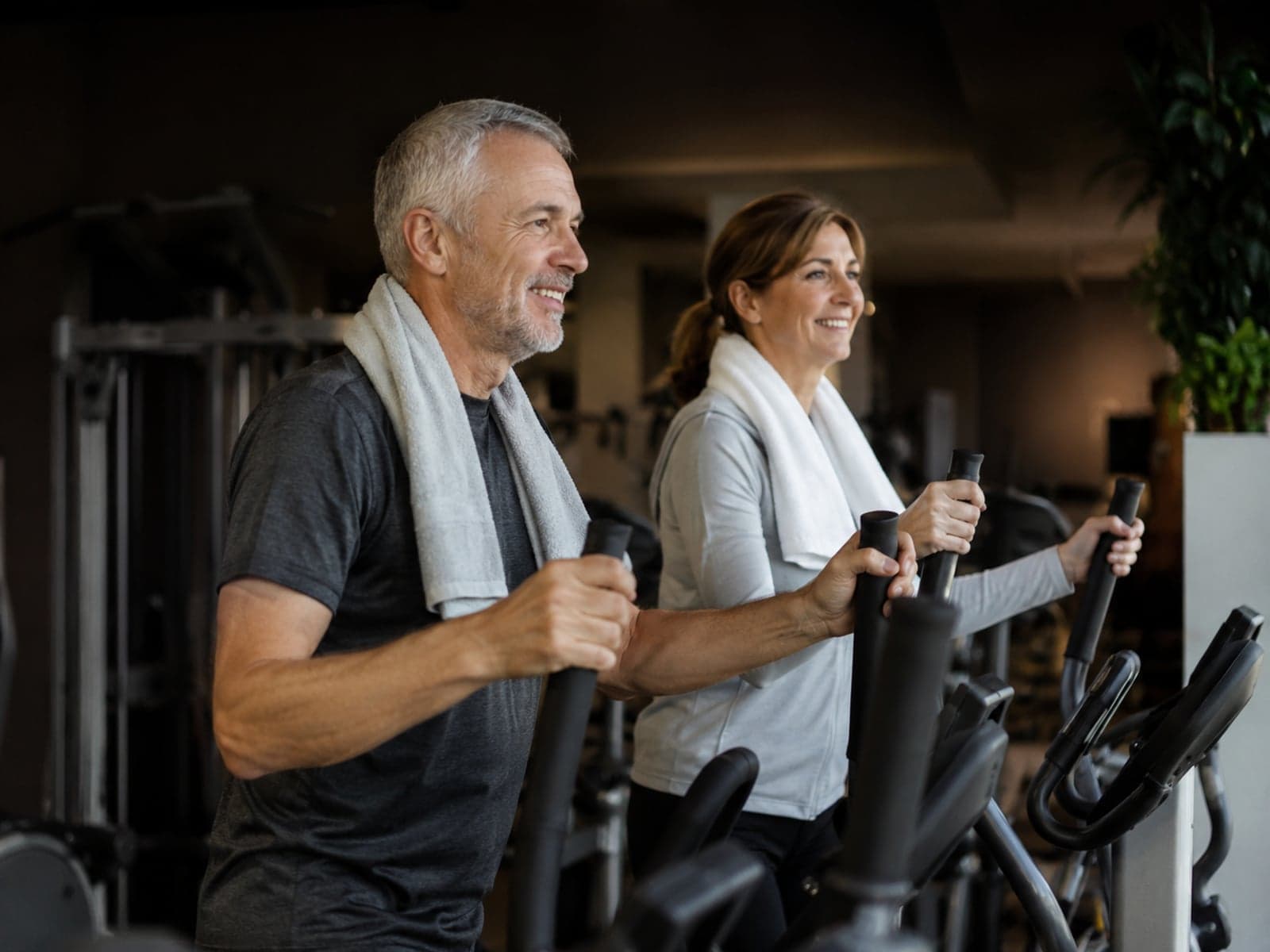 A man and a woman in their fifties smiling mid-workout on cross-trainers in a private studio